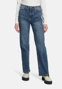 Jean en denim bleu avec une coupe décontractée, taille haute, poches avant et arrière, et décoloration subtile. Associé à des chaussures à semelle épaisse de couleur claire.