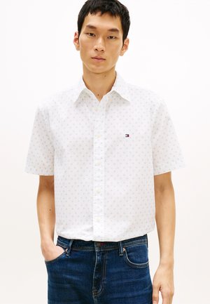 Νέος άνδρας που φοράει λευκό πουκάμισο με κοντά μανίκια και διακριτικό σχέδιο, καθώς και μπλε τζιν παντελόνι, στέκεται με το ένα χέρι στην τσέπη.