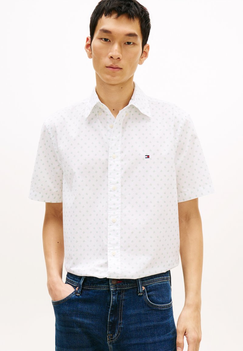 Νέος άνδρας που φοράει λευκό πουκάμισο με κοντά μανίκια και διακριτικό σχέδιο, καθώς και μπλε τζιν παντελόνι, στέκεται με το ένα χέρι στην τσέπη.