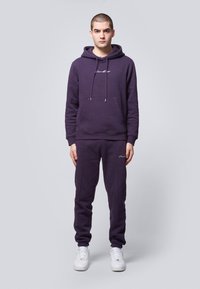 Purple cotton blend hoodie with front pocket and drawstrings, paired with matching joggers featuring side stripes, worn with white sneakers.