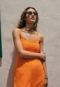 Combinaison sans manches orange avec broderie florale sur la poitrine, associée à des lunettes de soleil marron et à des colliers superposés, devant un mur clair.