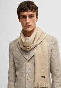 Vaalean beige neulehuivi, jossa on fringed yksityiskohtia, valahtaa rentoutuneesti vaalean beigeen teksturoituun takkiin. Huivin reunassa on huomaamaton logo.