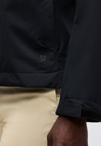 Κοντινό πλάνο ενός ατόμου που φοράει μαύρο μπουφάν με λογότυπο σε σχήμα καρδιάς κοντά στο τελείωμα και μπεζ παντελόνια, εμφανίζοντας τη μανσέτα και την τσέπη του μπουφάν.