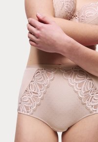 Beige hooggekraagde slip met stippenstof, kantafwerking en golvende randen. Heeft een zachte textuur met decoratieve elementen aan de zijkanten.
