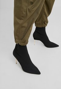 Bottines chaussettes noires côtelées à bouts pointus et talons fins dorés, portées avec un pantalon vert olive à cordon sur un fond gris clair.
