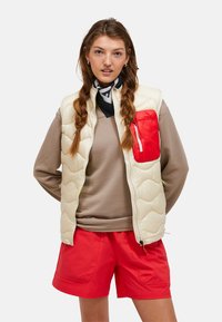 Gilet rembourré crème avec une poche poitrine rouge, porté par-dessus un haut marron texturé et un short rouge. Comprend une fermeture éclair et un col montant.