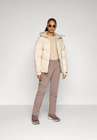 Lichtbeige donsjack, beige top en lichtbruine broek met zakken. Bruine wandelschoenen. Model draagt een zonnebril en staat tegen een neutrale achtergrond.