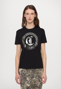 T-shirt en coton noir avec un logo circulaire en argent métallique présentant des motifs de serpents. Manches courtes et col rond. Associé à une jupe à imprimé serpent.