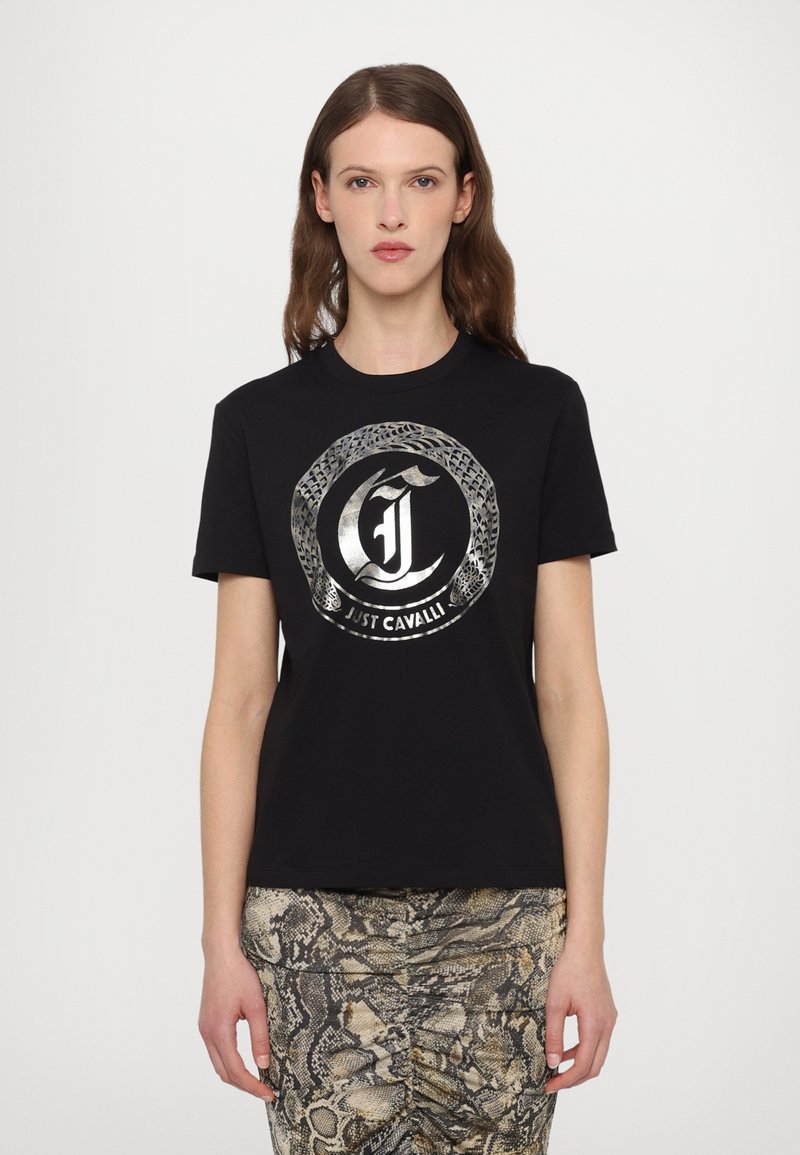 T-shirt en coton noir avec un logo circulaire en argent métallique présentant des motifs de serpents. Manches courtes et col rond. Associé à une jupe à imprimé serpent.