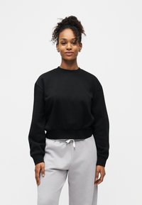 Νεαρή γυναίκα με σγουρά μαλλιά φορώντας μαύρο φούτερ και ανοιχτό γκρι φόρμα, στέκεται μπροστά σε απλό λευκό φόντο.