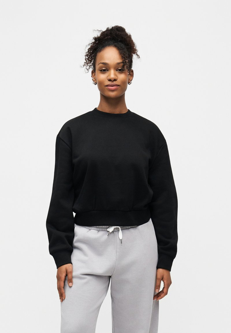 Νεαρή γυναίκα με σγουρά μαλλιά φορώντας μαύρο φούτερ και ανοιχτό γκρι φόρμα, στέκεται μπροστά σε απλό λευκό φόντο.