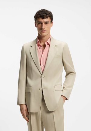 Mand iført en beige jakkesætjakke over en pink skjorte med knapper, står med den ene hånd i lommen mod en helt hvid baggrund.