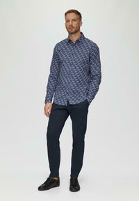 Niebieska, wzorzysta koszula zapinana na guziki z kołnierzykiem, w połączeniu z ciemnymi, dopasowanymi spodniami i czarnymi sneakersami. Koszula ma geometryczny wzór.