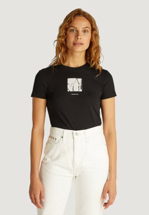 T-shirt nero a maniche corte con una grafica quadrata bianca che recita "CALVIN KLEIN" centrata sul petto, abbinato a un paio di jeans bianchi a vita alta.