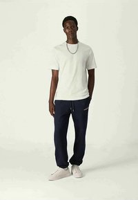 Man som bär en vit t-shirt, en silverhalsband, marinblå sweatpants och vita sneakers står mot en enkel bakgrund.