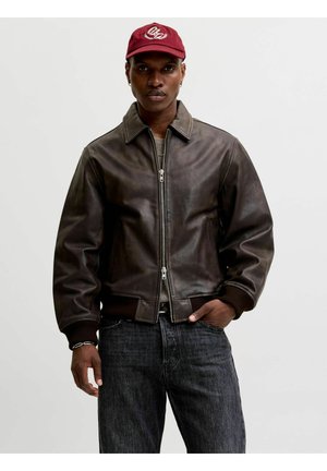 Homme portant une veste en cuir marron foncé zippée à mi-hauteur, un jean noir, une casquette de baseball rouge et des boucles d’oreilles argentées en forme d’anneau sur un fond uni.