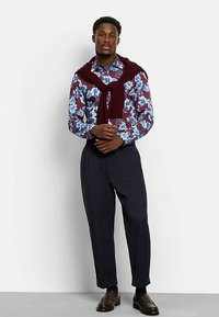 Blau- und burgunderrot gemustertes Hemd mit Blumenmuster, kombiniert mit dunklen, maßgeschneiderten Hosen. Ein bordeauxroter Pullover lässig über die Schultern gelegt, braune Schuhe.