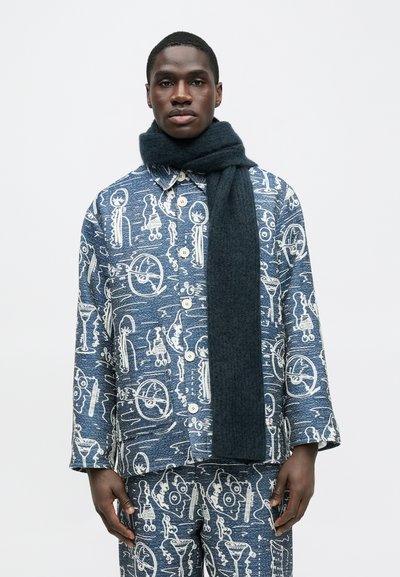 Μπλε πουκάμισο με λευκά αφηρημένα σχέδια, συνδυασμένο με σκούρο πλεκτό κασκόλ. Το ύφασμα φαίνεται υφασμένο με κουμπιά κλεισίματος.