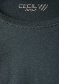 Tmavě zelené bavlněné tričko s kulatým výstřihem. Obsahuje stříbrný nápis "CECIL basic" a vzor srdcí. Hladká textura, minimalistický design.