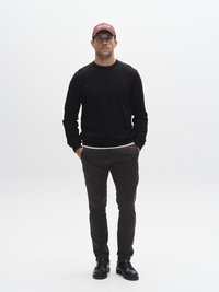 Schwarzer Langarm-Pullover, dunkelbraune Hose und schwarze Schuhe. Trägt eine burgunderrote Mütze mit Text, Hände in den Taschen, steht vor weißem Hintergrund.