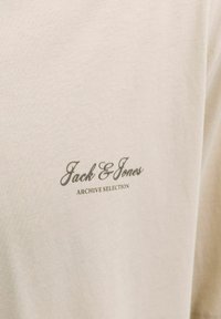 Μπεζ ύφασμα με τυπωμένο κείμενο "Jack & Jones" και μικρότερο κείμενο "ARCHIVE SELECTION".