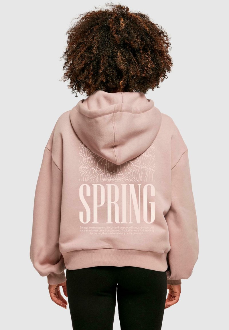 Svetloružová mikina s kapucňou, na chrbte s veľkým bielym textom "SPRING" a botanickým čiarovým umením. Mäkká textúra a voľný strih.