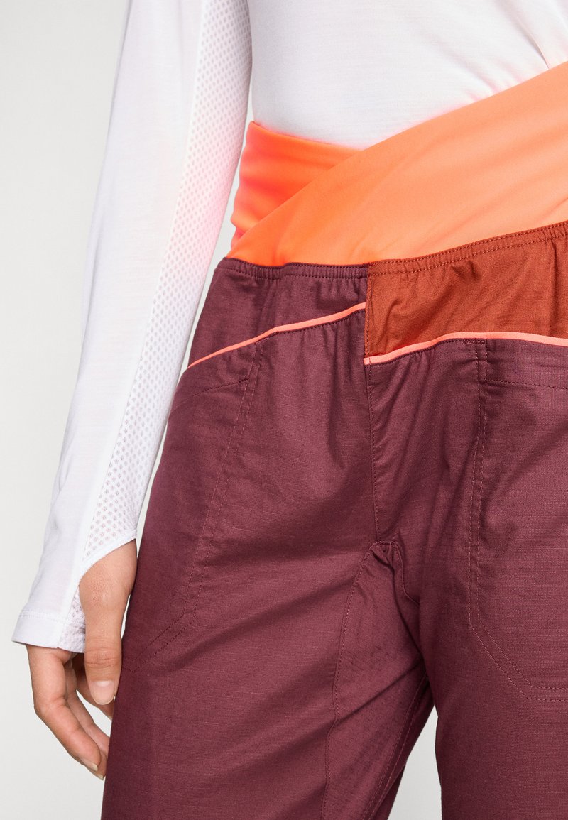Bordeauxfarbene Hose mit einem gemütlichen orangen Bündchen, das kontrastierende Nähte und Taschen aufweist. Das Oberteil ist weiß mit Netzdetails am Ärmel.