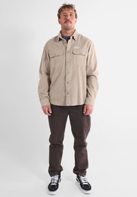 Camisa de pana beige con dos bolsillos en el pecho y cierre de botones, combinada con pantalones marrones y zapatillas negras con suelas blancas.