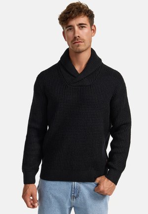 Svart genser med teksturert strikkemønster, sjalkrage, lange ermer og ribbestrikkede mansjetter og nederkant. Kombinert med lyseblå jeans.