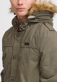 Parka vert olive avec une capuche en fourrure, fermeture éclair à l'avant, boutons-pression et un tissu texturé. Porte un écusson de marque sur la poitrine.