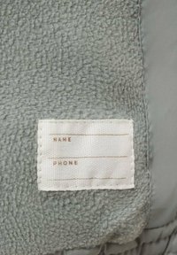 Szara tkanina o miękkiej, teksturowanej, przypominającej polar powierzchni. Biała metka z liniami "IMIĘ" i "NUMER TELEFONU" wyszytymi jasnobrązową nicią.