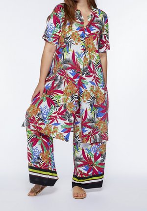 Femme portant une tunique à motif floral coloré et un pantalon large assorti avec des sandales, debout sur un fond blanc.