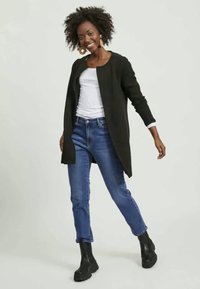 Χαμογελαστή γυναίκα με σγουρά μαλλιά, φορώντας μεγάλα στρογγυλά σκουλαρίκια, μαύρο ανοιχτό τζάκετ, λευκό πουκάμισο, μπλε τζιν και μαύρες μπότες ankle, ποζάροντας πλευρικά.