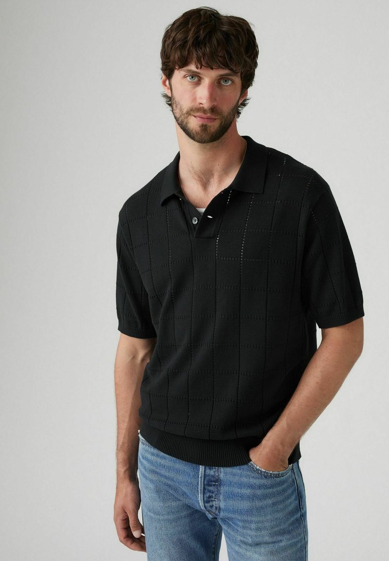 Schwarzes gestricktes Poloshirt mit einem strukturierten Schachbrettmuster, kurzen Ärmeln und einem klassischen Kragen, kombiniert mit hellblauen Jeans.