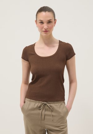 Junge Frau mit zurückgebundenem Haar, die ein braunes Kurzarmoberteil und beige Hose mit Kordelzug trägt, steht mit den Händen in den Taschen.
