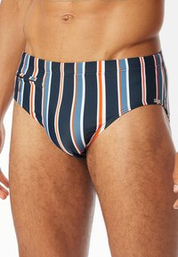 Randig badbralla i marinblått med vertikala röd, vit och blå ränder. Har en ficka med dragkedja och en slät, elastisk tygkänsla.