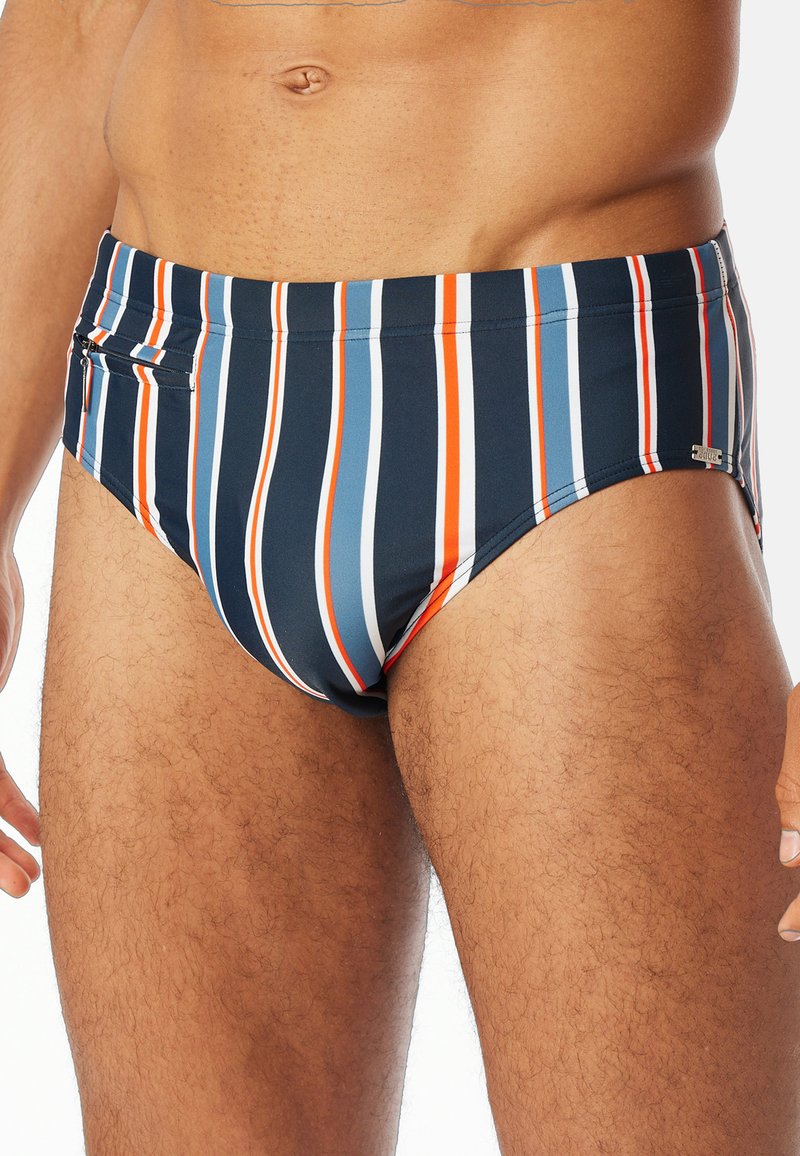 Randig badbralla i marinblått med vertikala röd, vit och blå ränder. Har en ficka med dragkedja och en slät, elastisk tygkänsla.