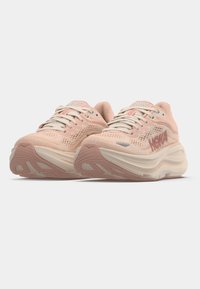 Ζεύγος ροζ και μπεζ αθλητικών παπουτσιών HOKA με παχύ μαξιλαρωτό πάτο και αναπνεύσιμες διχτυωτές επάνω επιφάνειες, εμφανιζόμενα από χαμηλή εμπροσθο πλευρά.