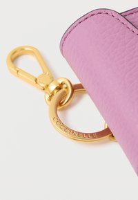 Etui pentru chei din piele texturată roz cu accesorii aurii, prevăzut cu un inel rotund cu embossing "COCCINELLE" și o carabină pivotantă.