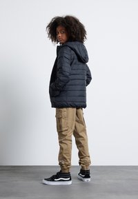 Černá péřová bunda s kapucí, béžové cargo kalhoty a černé tenisky Vans. Hladká textura, výrazný nadýchaný design a elastické manžety.