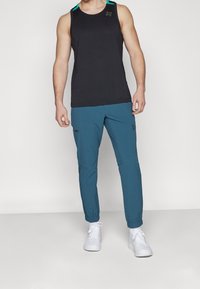 Camiseta sin mangas negra con detalles en verde azulado y logo, combinada con pantalones jogger ajustados en verde azulado con bolsillos y puños elásticos, llevados con zapatillas blancas.