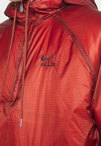 Röd nylon vindjacka med huva, med en texturerad yta, svart "Nike Air"-logga och snörning i halsen.