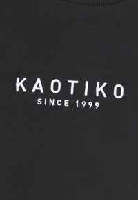Tissu noir avec texte brodé blanc "KAOTIKO" au-dessus d'un texte plus petit "SINCE 1999" centré.