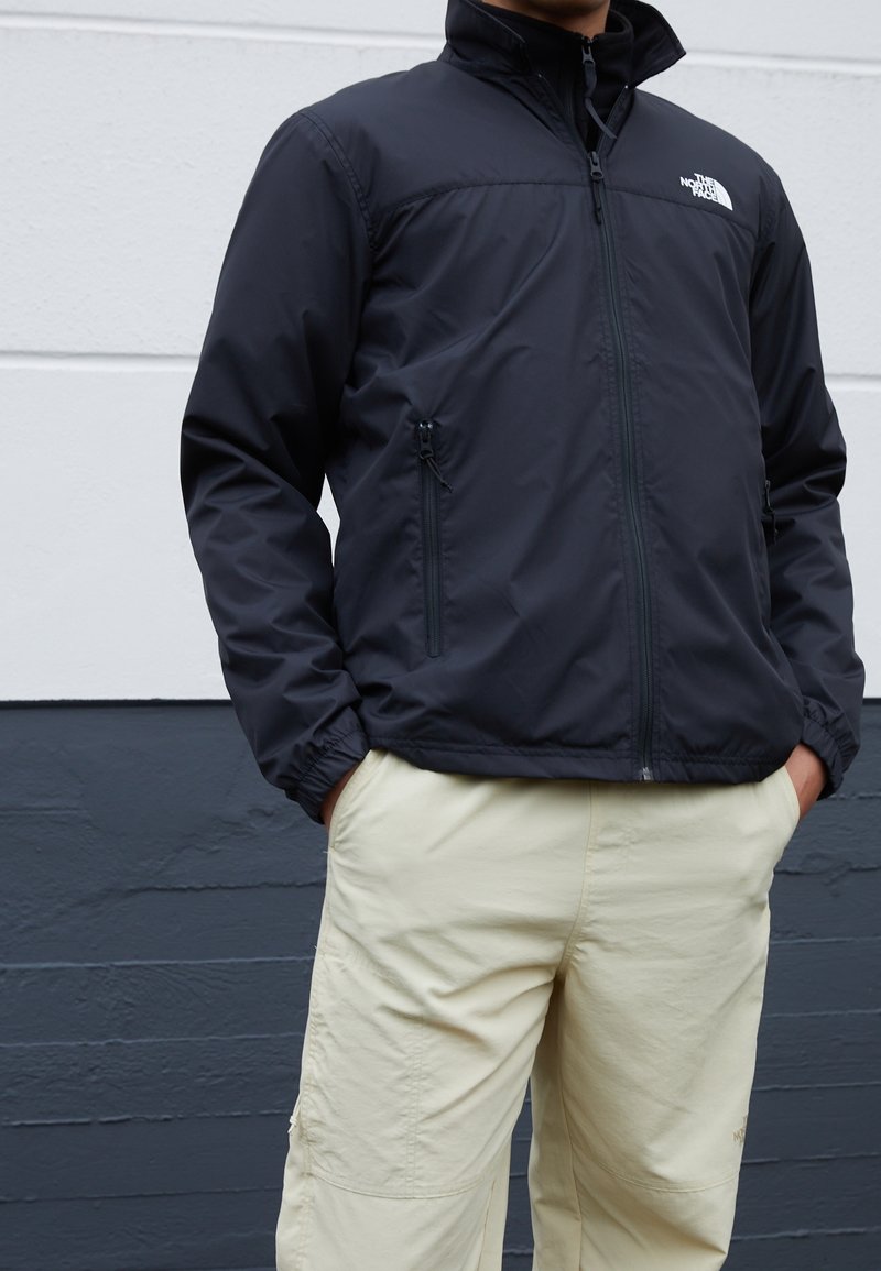 Personne portant une veste North Face noire avec des poches zippées et un pantalon beige clair, debout contre un mur gris bicolore.