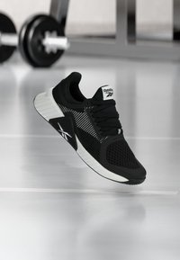 Αθλητικό παπούτσι Reebok σε ασπρόμαυρο, κρεμασμένο στον αέρα σε γυμναστήριο, με θαμπά βάρη στο φόντο.