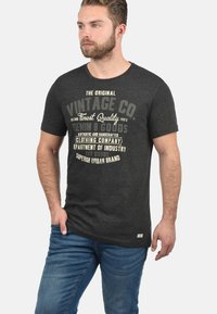 Camiseta negra con texto gráfico de estilo vintage en crema y gris. Manga corta, material de algodón, ajuste casual, combinada con vaqueros azules.