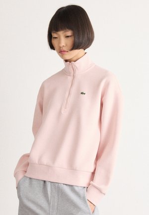 Jeune femme aux cheveux noirs courts, portant un sweatshirt à zip quart rose pâle et un pantalon gris, se tenant les mains dans les poches.