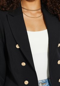 Blazer noir ajust é avec des boutons dorés, présentant un design à double boutonnage. Colliers en chaîne dorée superposés sur un haut blanc.