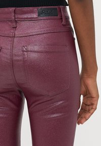 Jeans skinny bordeaux com um acabamento brilhante e texturizado, apresentando dois bolsos traseiros e um rótulo preto na cintura.
