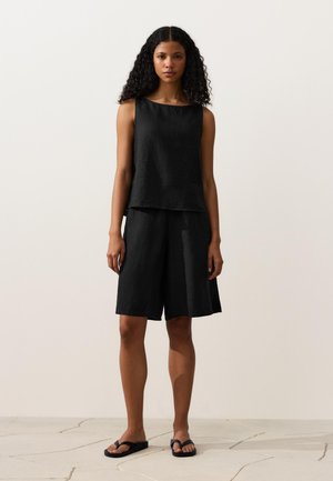 Jonge vrouw met krullend haar, gekleed in een zwarte mouwloze top, zwarte knielange shorts en zwarte slippers, staand op een lichte vloer tegen een effen muur.